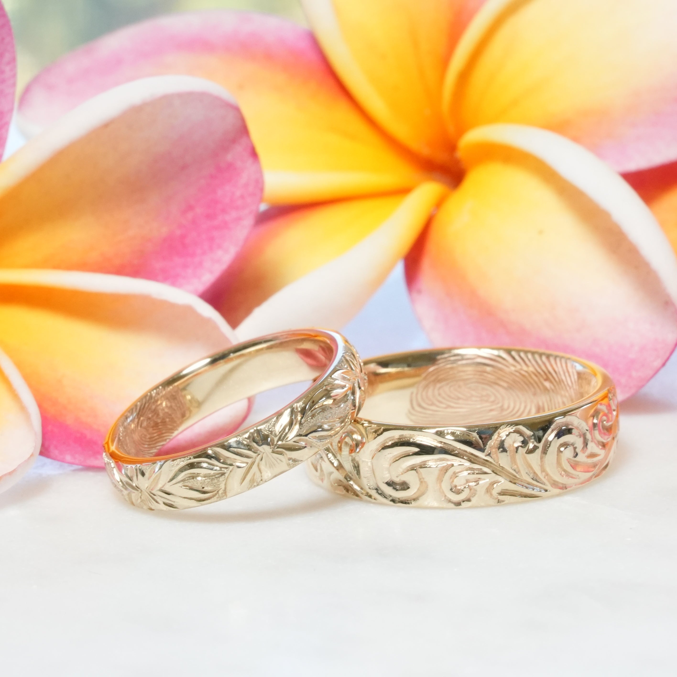 Two gold rings with intricate designs on a white surface with pink and yellow flowers in the background.