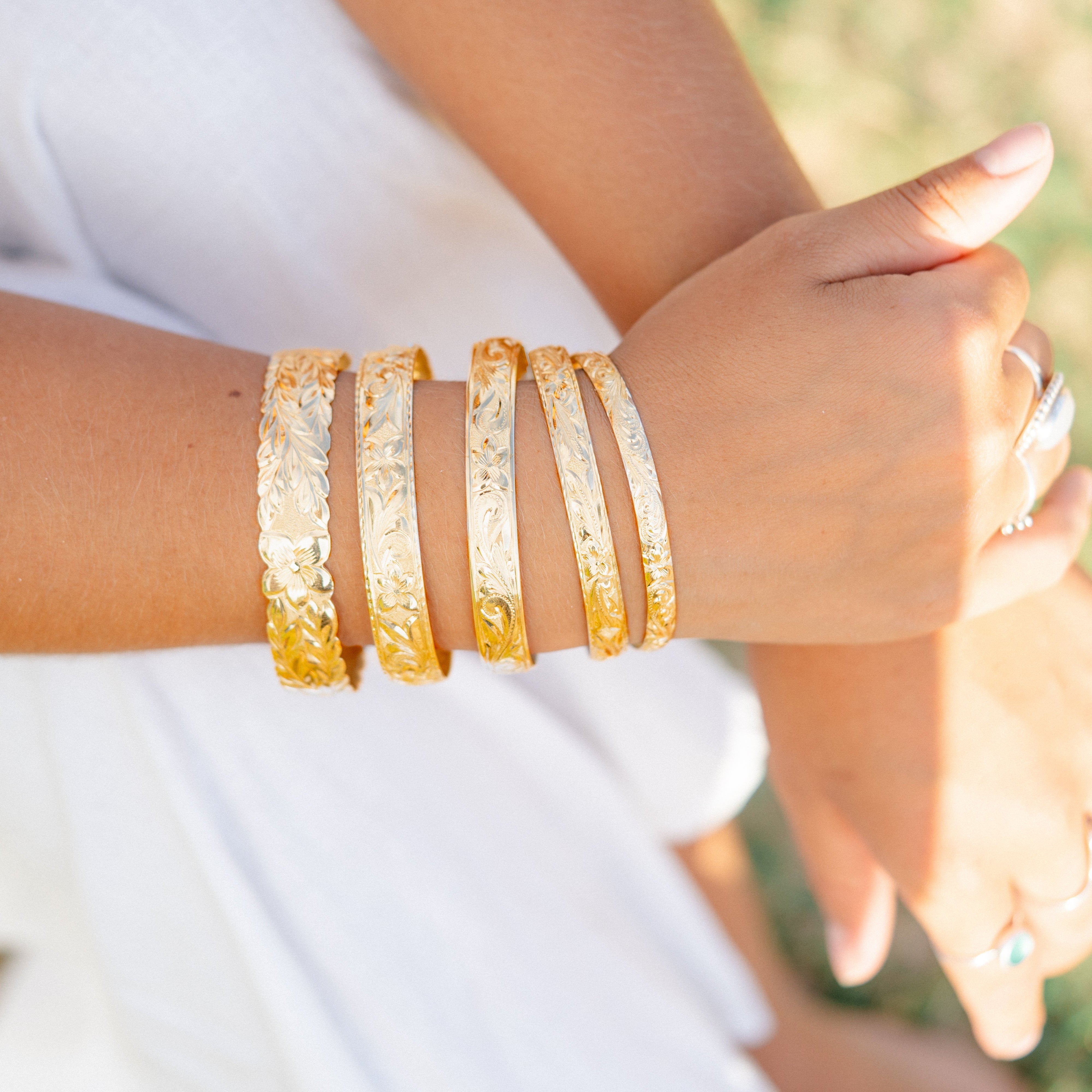 Close-up of a person's wrist wearing multiple gold hawaiian heirloom bracelets with a blurred natural background on Kauai Hawaii